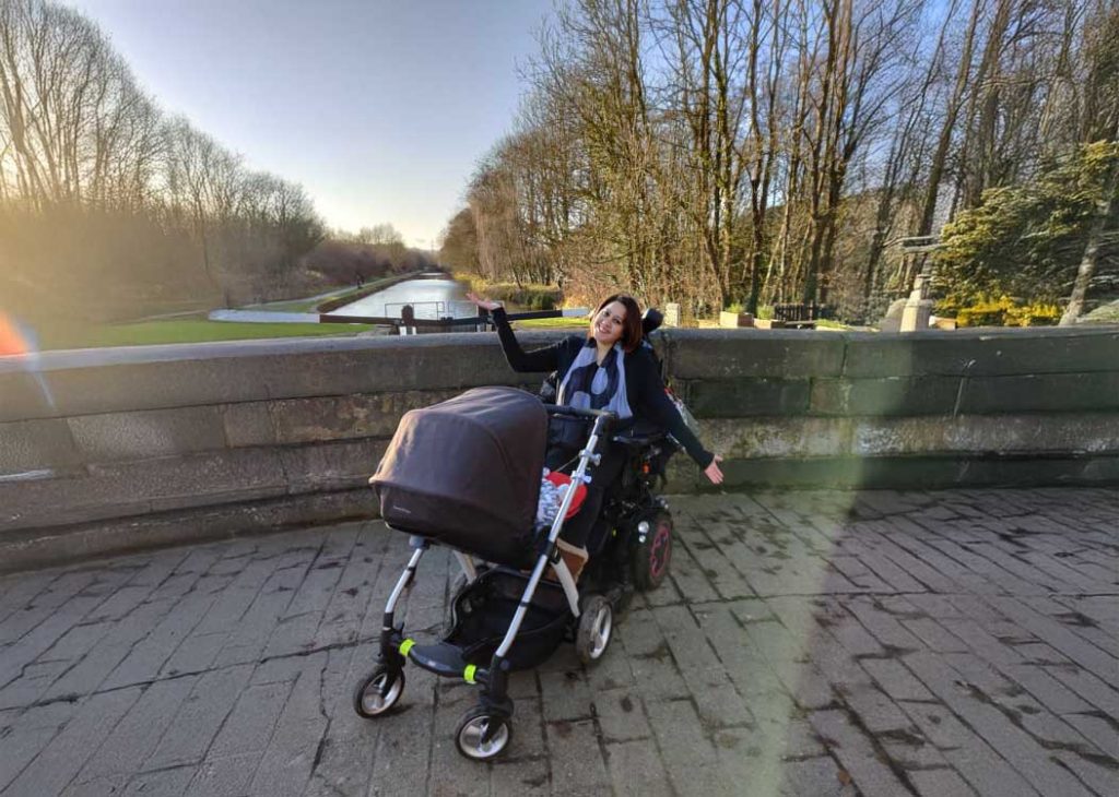 Aisha, smiling and sitting in a power wheelchair with a baby stroller attached, gestures toward a scenic canal and trees on a sunny day.