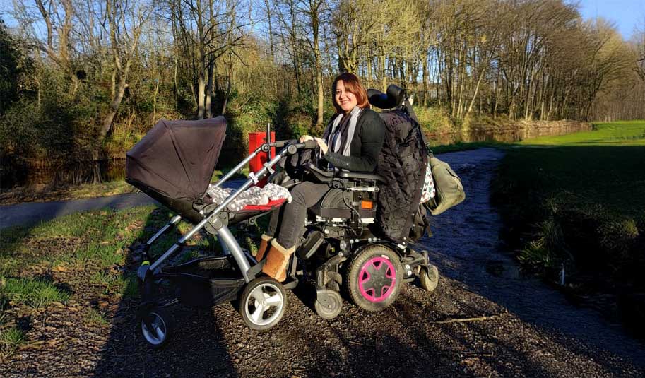 Aisha, smiling in her power wheelchair with a baby stroller, on a path surrounded by trees and grass on a sunny day.