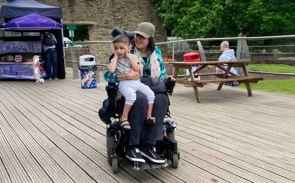 Aisha, smiling in her power wheelchair while holding a young boy on her lap, at an outdoor public space with wooden decking and picnic tables.