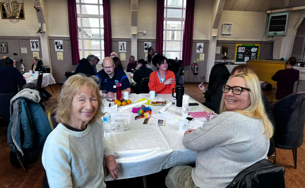 Two women smiling at a table during a volunteer training session, with other participants seated at tables in a community hall behind them.